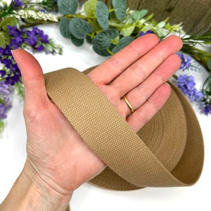 May include: A close-up of a roll of tan cotton webbing. The webbing is 1 inch wide and has a smooth, woven texture.