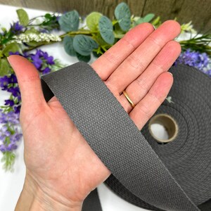 May include: A roll of dark gray cotton webbing, approximately 1 inch wide, is held in a hand. The webbing is woven with a textured pattern.