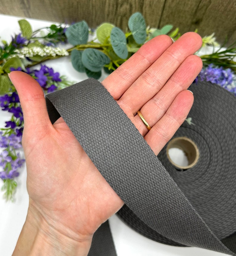 May include: A roll of dark gray cotton webbing, approximately 1 inch wide, is held in a hand. The webbing is woven with a textured pattern.