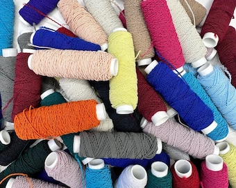 Op de afbeelding: Een verzameling van talloze klossen garen in een grote verscheidenheid aan kleuren, waaronder blauw, oranje, geel, roze, rood, grijs, wit en groen. Elke klos heeft een witte plastic kern en is omwikkeld met getextureerd, meerdraads garen.