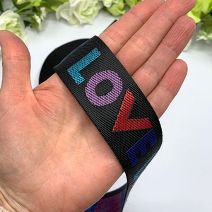 May include: Black elastic band with the word "LOVE" printed in colorful letters. The letters are outlined in a contrasting color. The band is held in a hand.