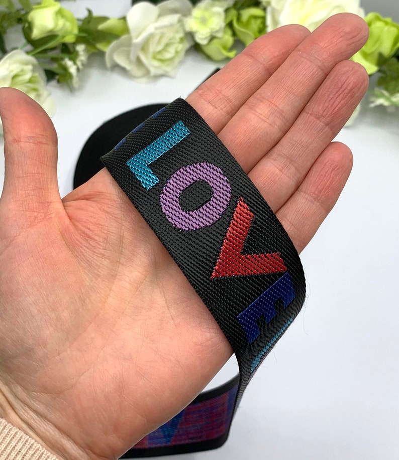 May include: Black elastic band with the word "LOVE" printed in colorful letters. The letters are outlined in a contrasting color. The band is held in a hand.