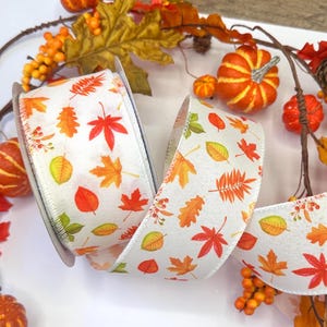 May include: A white fabric ribbon featuring a repeating pattern of colorful autumn leaves in shades of orange, red, yellow, and green. The ribbon has a wired edge and is displayed in a loose coil, showcasing the detailed botanical print.