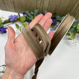 K&ouml;nnte beinhalten: Zwei Str&auml;nge braunes Samtband, einer in einer etwas helleren Farbe, werden in einer Hand gehalten. Das Band ist weich und hat einen dezenten Glanz.
