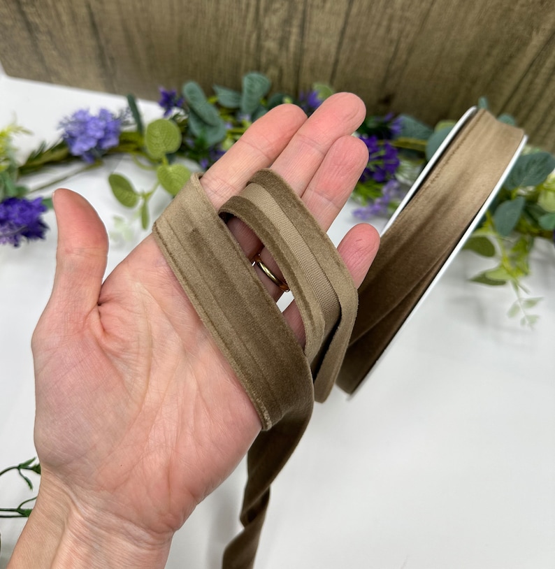 K&ouml;nnte beinhalten: Zwei Str&auml;nge braunes Samtband, einer in einer etwas helleren Farbe, werden in einer Hand gehalten. Das Band ist weich und hat einen dezenten Glanz.