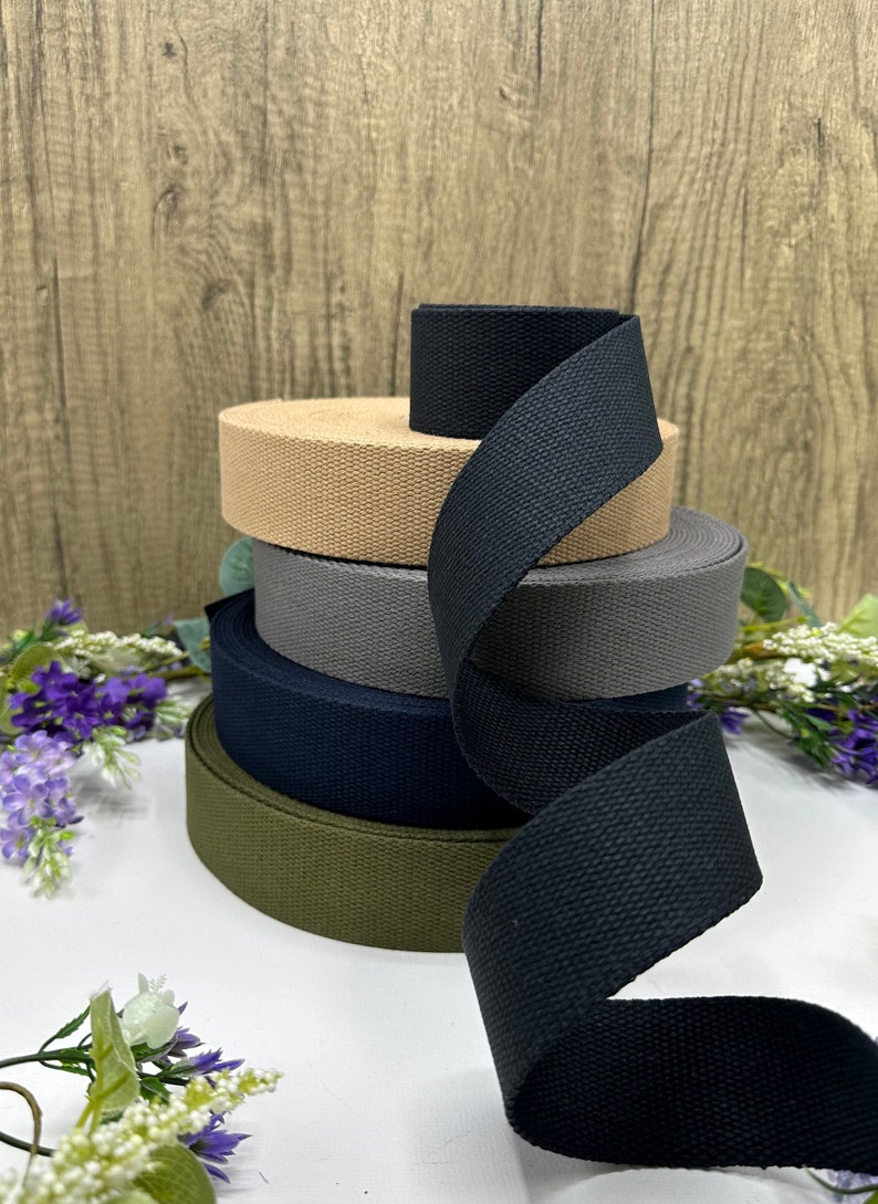 May include: A stack of five cotton webbing straps in different colors, including black, khaki, dark green, navy blue, and gray. The straps are rolled up and stacked on top of each other.