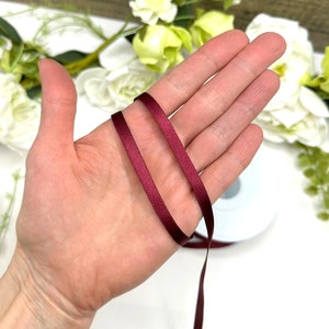 May include: Two strands of burgundy satin ribbon, approximately 1/4 inch wide, are shown wrapped around a hand.