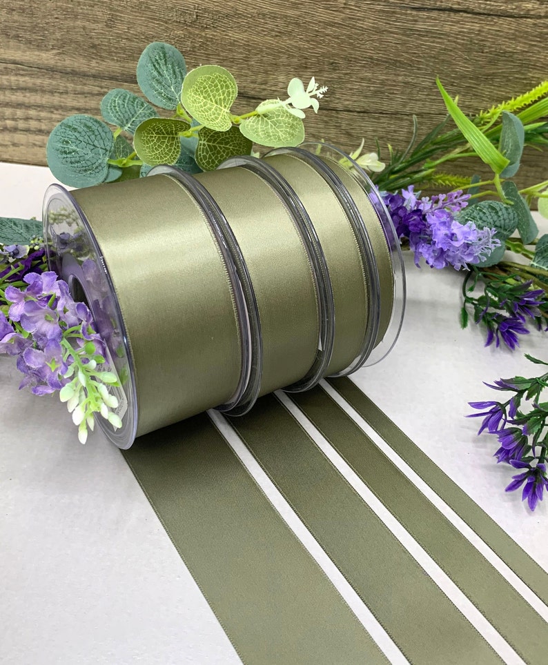 May include: Four spools of olive green satin ribbon. The ribbon is wound around clear plastic spools. The ribbon is lying on a white surface.