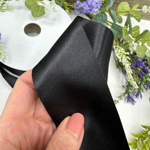 May include: A roll of black satin ribbon. The ribbon is folded in half and held in a hand. The ribbon is smooth and shiny.