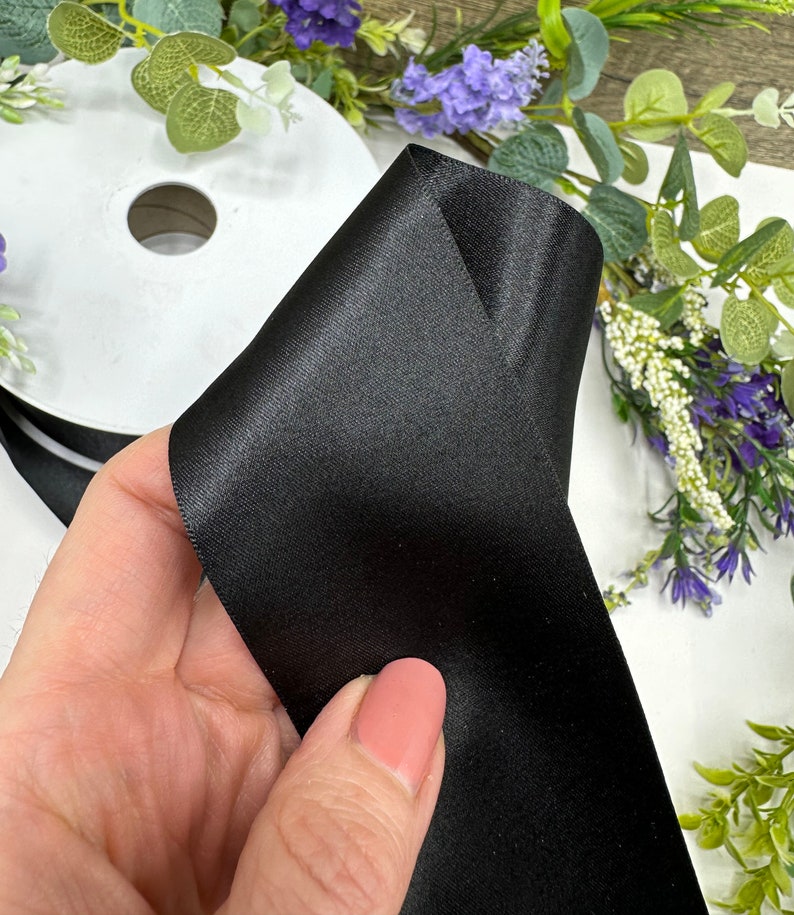 May include: A roll of black satin ribbon. The ribbon is folded in half and held in a hand. The ribbon is smooth and shiny.