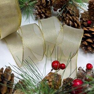 Puede incluir: Un rollo de cinta transparente color crema con ribete dorado, parcialmente desenrollado. La cinta est&aacute; rodeada de pi&ntilde;as, bayas rojas y agujas de pino verdes, creando un arreglo festivo navide&ntilde;o.