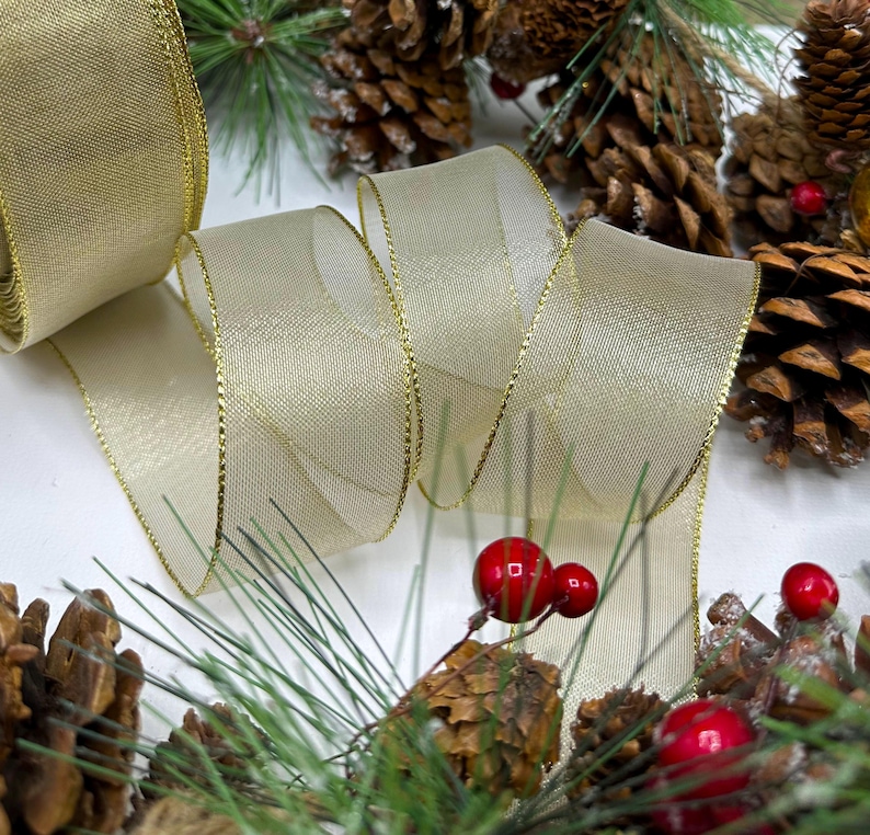 Puede incluir: Un rollo de cinta transparente color crema con ribete dorado, parcialmente desenrollado. La cinta est&aacute; rodeada de pi&ntilde;as, bayas rojas y agujas de pino verdes, creando un arreglo festivo navide&ntilde;o.