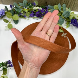 May include: A spool of brown woven cotton webbing. The webbing is a solid, warm brown color and appears to be tightly woven. The webbing is being held in a hand, with a gold ring on the finger. The background includes greenery and flowers.