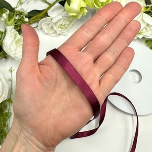 May include: A spool of burgundy satin ribbon, approximately 1/4 inch wide, is wrapped around a hand. The ribbon is smooth and shiny.