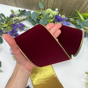 Pode incluir: Um rolo de fita de veludo bordô com uma borda dourada. A fita é segurada em uma mão, mostrando sua cor rica e textura. A borda dourada adiciona um toque de elegância, adequada para vários projetos de artesanato.