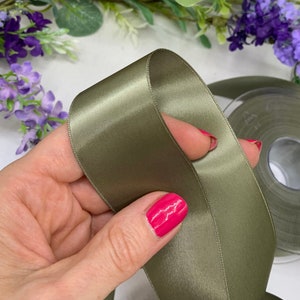 May include: A roll of sage green satin ribbon. The ribbon is folded in half and held in a hand.