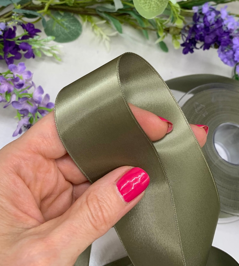 May include: A roll of sage green satin ribbon. The ribbon is folded in half and held in a hand.