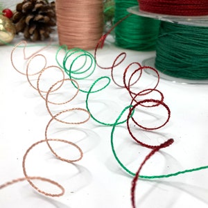May include: Close-up of decorative spools of thread in shades of green, red, and copper. The thread is arranged in swirling patterns, suggesting a festive or craft theme. Additional spools of thread are visible in the background.