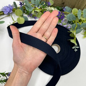 May include: A roll of navy blue cotton webbing is held in a hand. The webbing is a flat, woven tape, and is unspooled from a cardboard core. The background includes greenery and purple flowers.