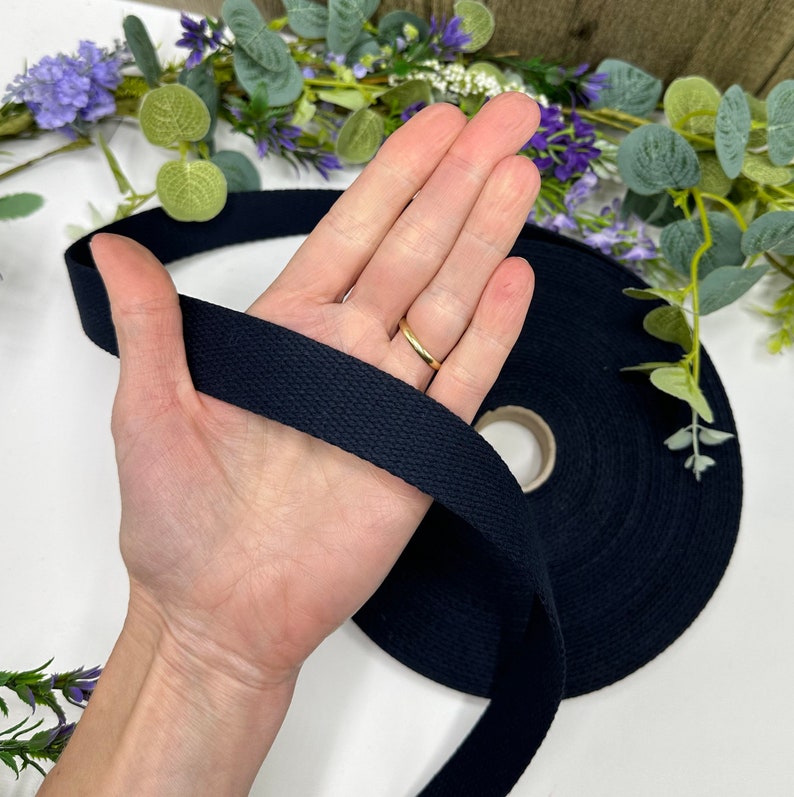 May include: A roll of navy blue cotton webbing is held in a hand. The webbing is a flat, woven tape, and is unspooled from a cardboard core. The background includes greenery and purple flowers.