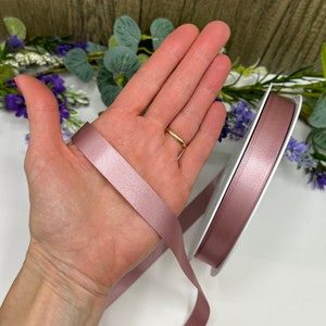 May include: A roll of dusty rose satin ribbon, approximately 2.5 cm wide, is shown on a white surface. A hand is holding a length of the ribbon, showing the sheen and texture of the fabric.