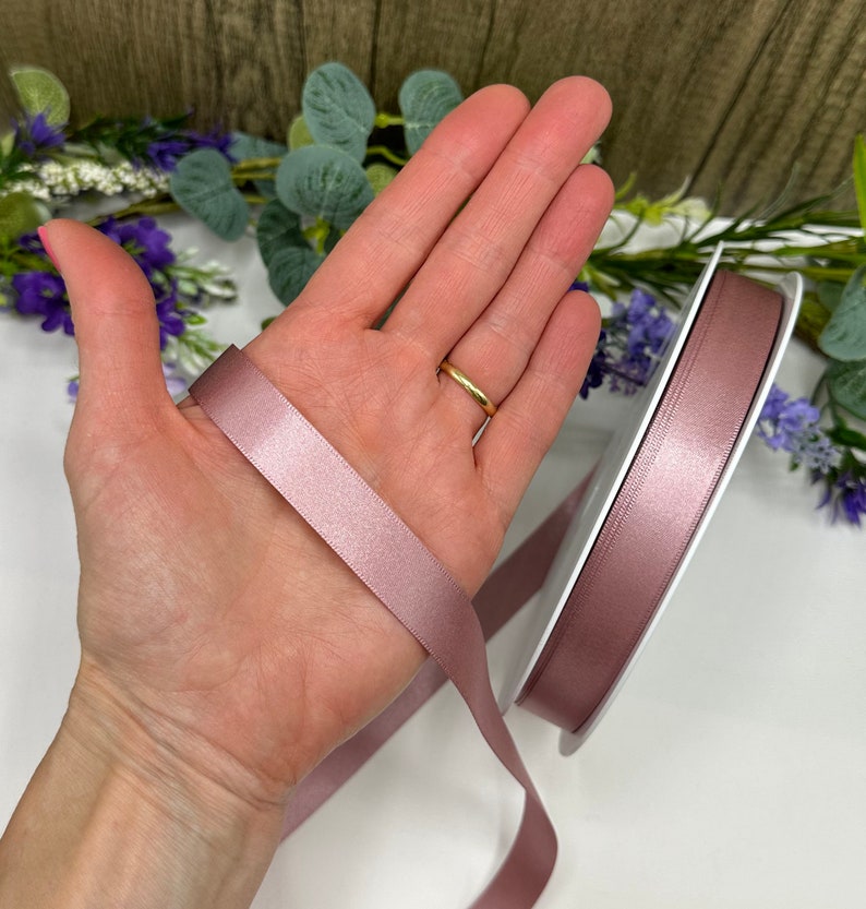 May include: A roll of dusty rose satin ribbon, approximately 2.5 cm wide, is shown on a white surface. A hand is holding a length of the ribbon, showing the sheen and texture of the fabric.