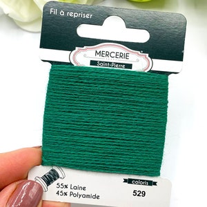 May include: A spool of green embroidery floss with the label "Mercerie Saint-Pierre" and the text "Fil &agrave; repriser" on the top. The label also includes the text "55% Laine 45% Polyamide" and "coloris 529".