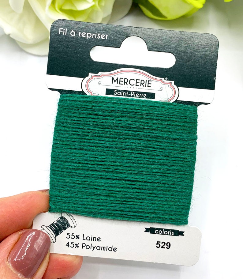 May include: A spool of green embroidery floss with the label "Mercerie Saint-Pierre" and the text "Fil &agrave; repriser" on the top. The label also includes the text "55% Laine 45% Polyamide" and "coloris 529".