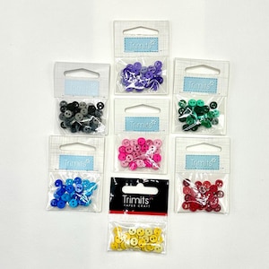 May include: A selection of colourful buttons in small plastic bags. Colours include purple, black, green, pink, blue, red, and yellow. Each bag is labelled with the brand name "Trimmits". The yellow buttons have a smiley face design.