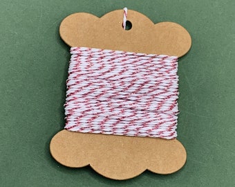 May include: A spool of white twine with a thin red and silver glitter thread running through it. The twine is wound around a brown cardboard spool with a scalloped edge.
