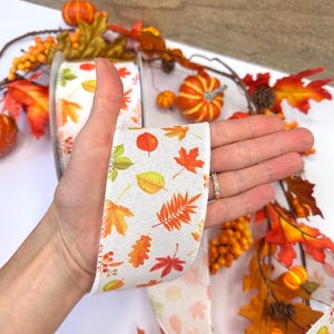 May include: A white fabric ribbon featuring a repeating pattern of colorful autumn leaves in shades of orange, yellow, red, and green. The ribbon has a textured weave and is displayed against a background of artificial fall foliage.