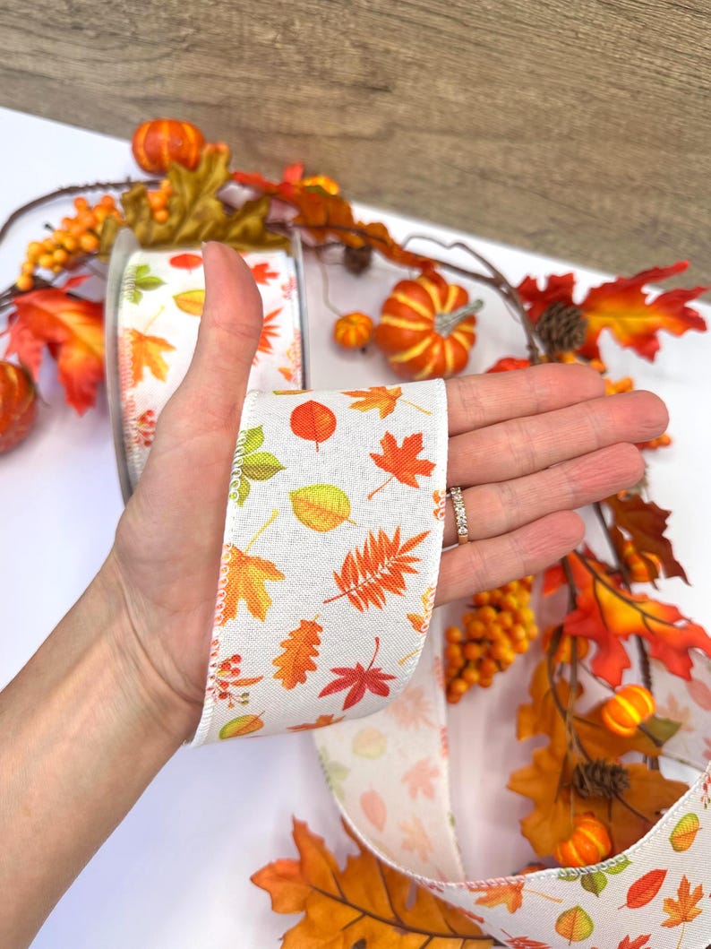 May include: A white fabric ribbon featuring a repeating pattern of colorful autumn leaves in shades of orange, yellow, red, and green. The ribbon has a textured weave and is displayed against a background of artificial fall foliage.