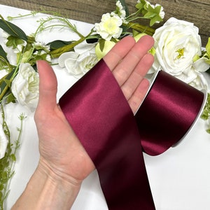May include: A roll of deep burgundy satin ribbon. The ribbon is shown on a hand, with the ends of the ribbon folded over.