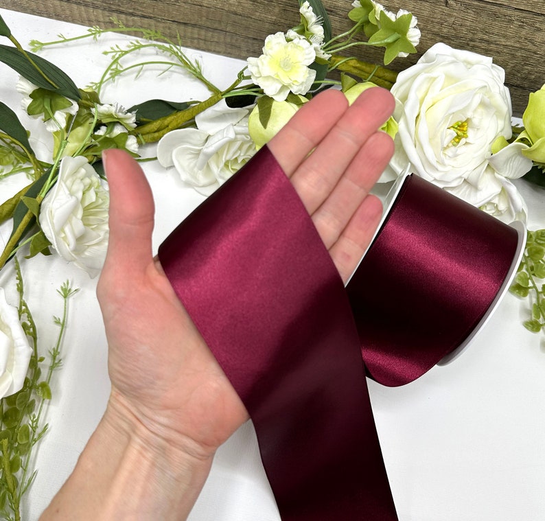 May include: A roll of deep burgundy satin ribbon. The ribbon is shown on a hand, with the ends of the ribbon folded over.