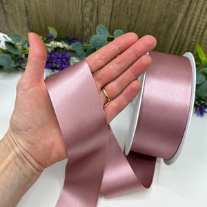 May include: A roll of dusty rose satin ribbon. The ribbon is shown on a white spool and in the hand of a person.