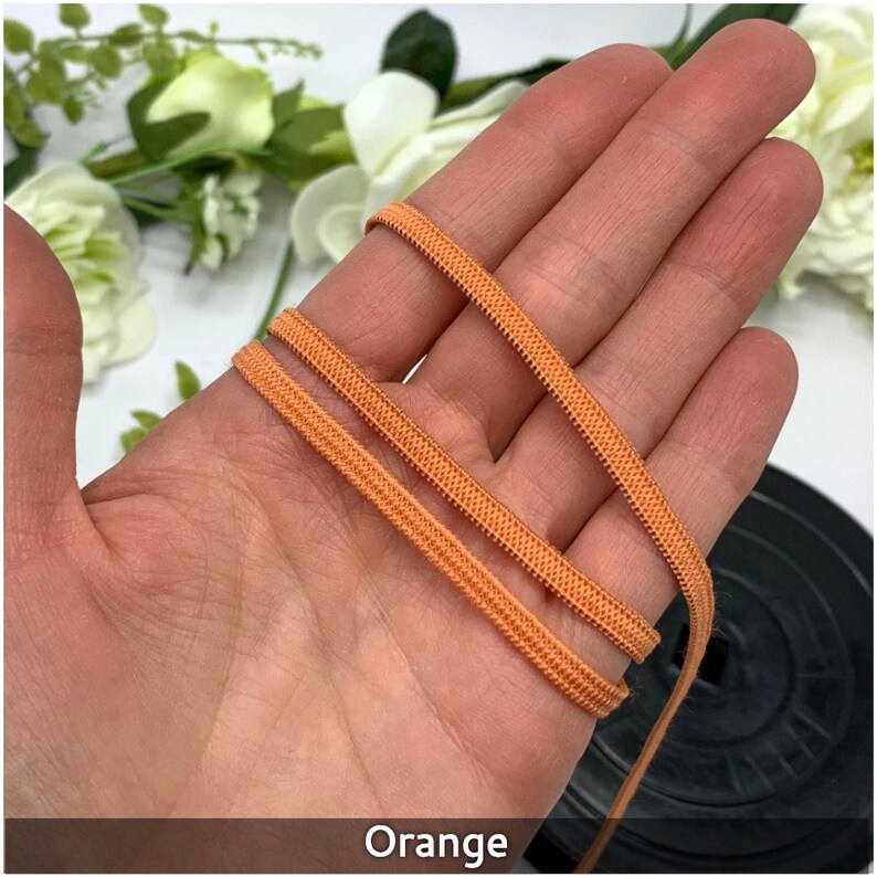 May include: Orange elastic cord, a type of stretch fabric, is shown in a close-up image. The cord is folded in half and held in a hand. The cord is a bright orange color.