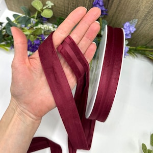 K&ouml;nnte beinhalten: Burgunderfarbenes Samtband mit Satin-Finish. Das Band ist etwa 2,5 cm breit und wird auf einer Spule gezeigt.
