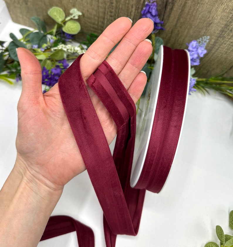K&ouml;nnte beinhalten: Burgunderfarbenes Samtband mit Satin-Finish. Das Band ist etwa 2,5 cm breit und wird auf einer Spule gezeigt.