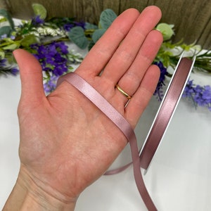 May include: A roll of dusty rose satin ribbon, approximately 6mm wide, is shown on a white surface. The ribbon is held in a hand, showing the width and colour of the ribbon.