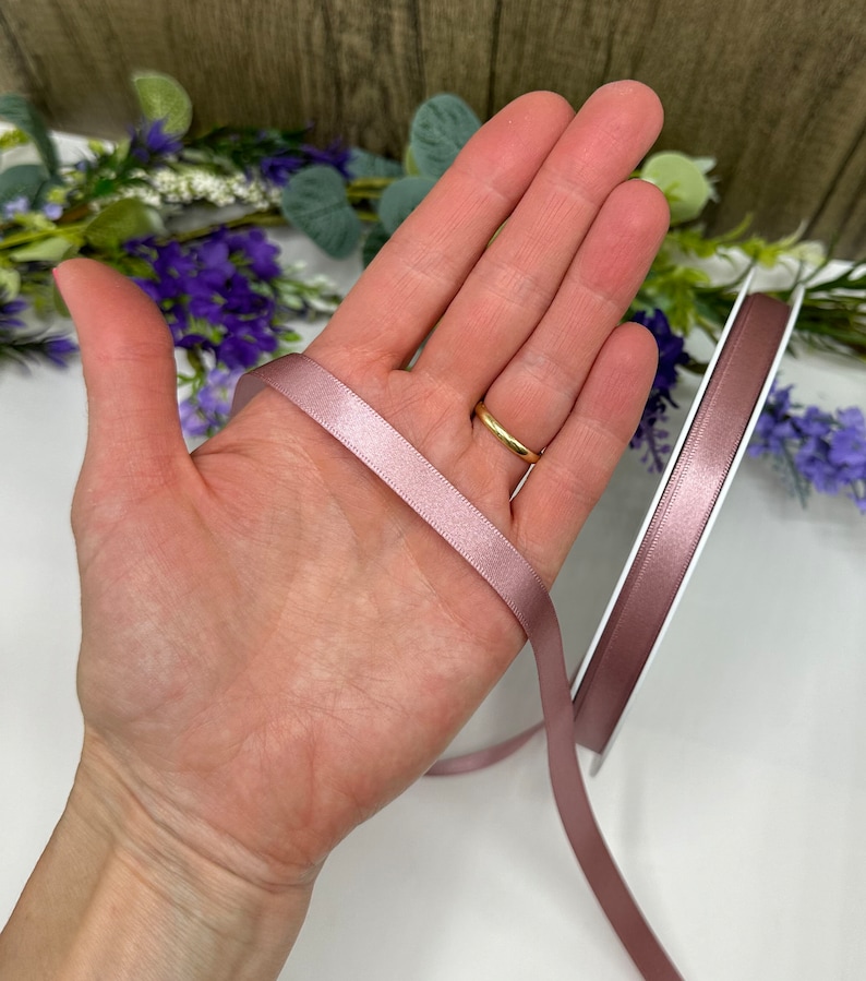 May include: A roll of dusty rose satin ribbon, approximately 6mm wide, is shown on a white surface. The ribbon is held in a hand, showing the width and colour of the ribbon.