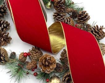 Op de afbeelding: Een breed rood fluwelen lint met een metallic gouden achterkant en goudkleurige randen, gedrapeerd over een decoratieve krans met bruine dennenappels, groene dennennaalden, kleine rode bessen en een enkele gouden kerstbal.