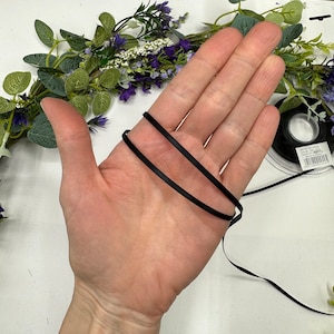 May include: A close-up of a hand holding two strands of black satin ribbon. The ribbon is approximately 1/4 inch wide.