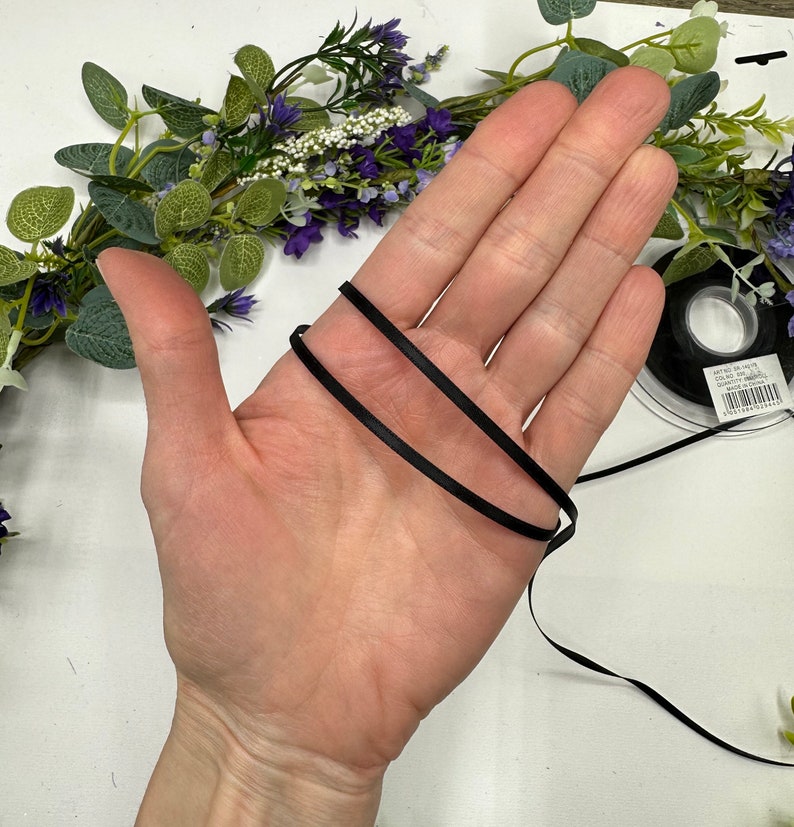 May include: A close-up of a hand holding two strands of black satin ribbon. The ribbon is approximately 1/4 inch wide.