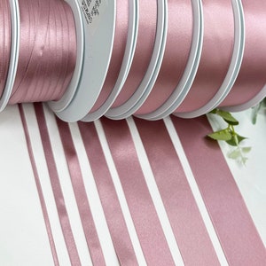 May include: Assortment of dusty rose satin ribbon spools in various widths. The ribbon is wound around white spools and laid out on a white surface.