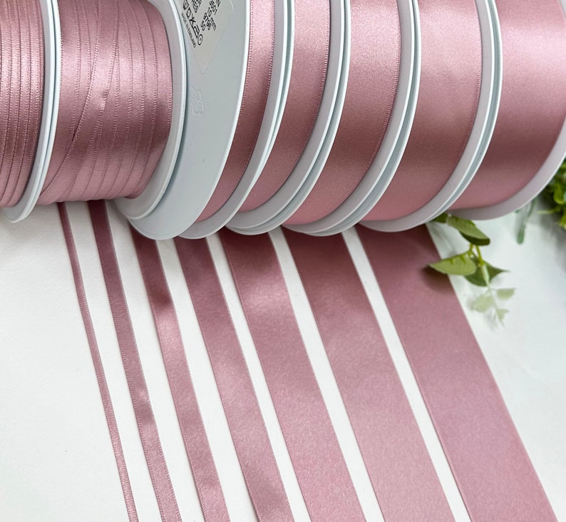 May include: Assortment of dusty rose satin ribbon spools in various widths. The ribbon is wound around white spools and laid out on a white surface.