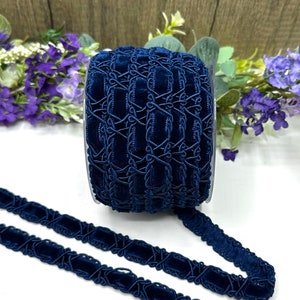 May include: A spool of navy blue velvet ribbon with an intricate, geometric design. The ribbon is displayed against a backdrop of wooden planks and purple flowers, showcasing its rich color and texture. The ribbon is approximately 1 inch wide.
