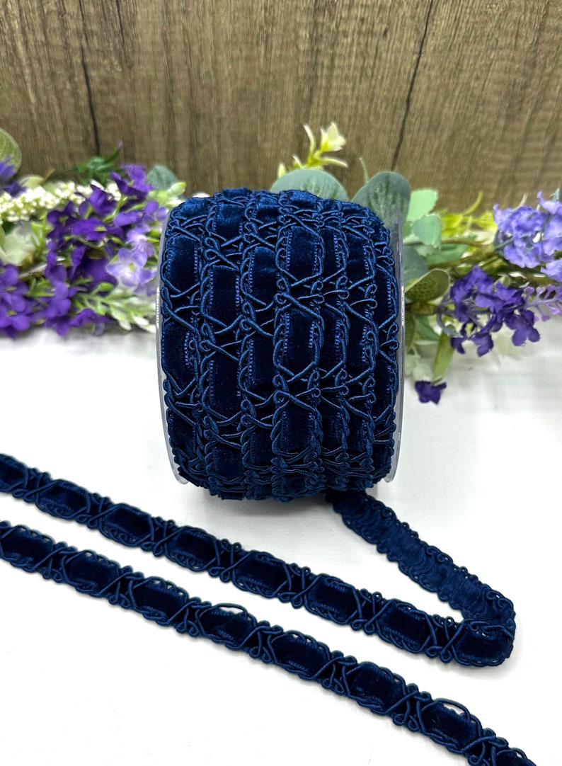 May include: A spool of navy blue velvet ribbon with an intricate, geometric design. The ribbon is displayed against a backdrop of wooden planks and purple flowers, showcasing its rich color and texture. The ribbon is approximately 1 inch wide.