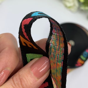 May include: Black woven ribbon with colorful rainbow threads and a repeating pattern of letters. The ribbon is folded in half, showing the inside and outside of the ribbon.