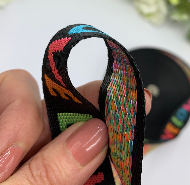 May include: Black woven ribbon with colorful rainbow threads and a repeating pattern of letters. The ribbon is folded in half, showing the inside and outside of the ribbon.