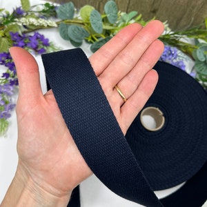 May include: A roll of black cotton webbing, approximately 1 inch wide, is shown in the image. The webbing is a woven fabric with a smooth texture. The webbing is being held in a person's hand.